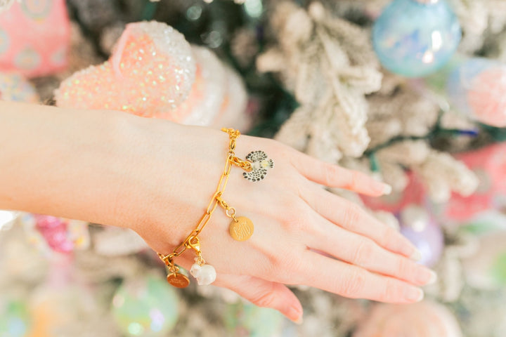 A gold charm bracelet with a snowflake and round disc charms, worn on a person's wrist against a soft-focus Christmas tree background.