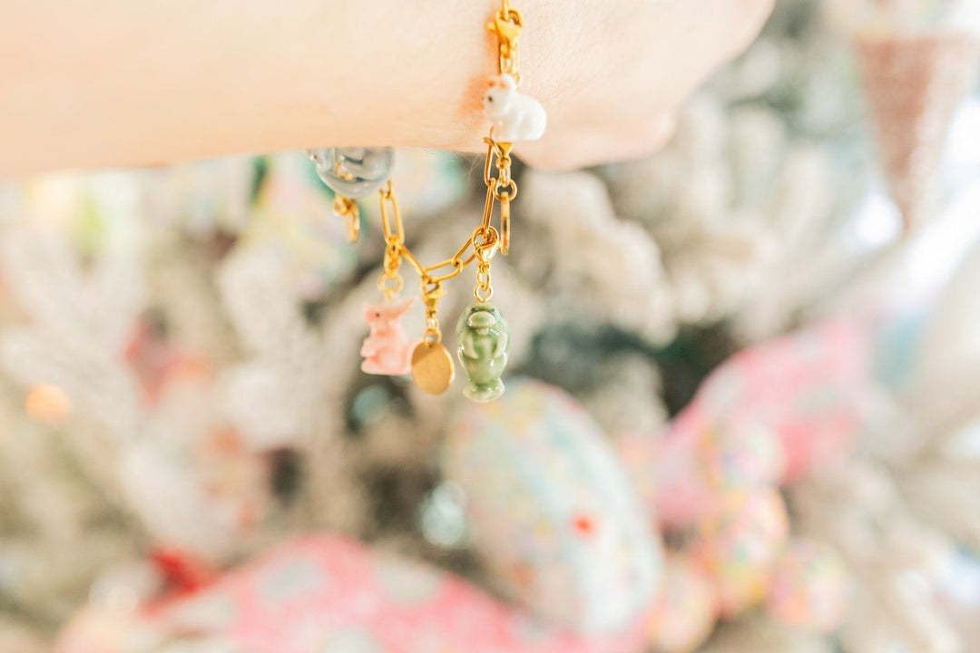 A close-up lifestyle shot of a delicate gold chain bracelet worn on a wrist, featuring pastel charms including a pink bunny, a green frog, and white pearls, set against a soft, blurred background.
