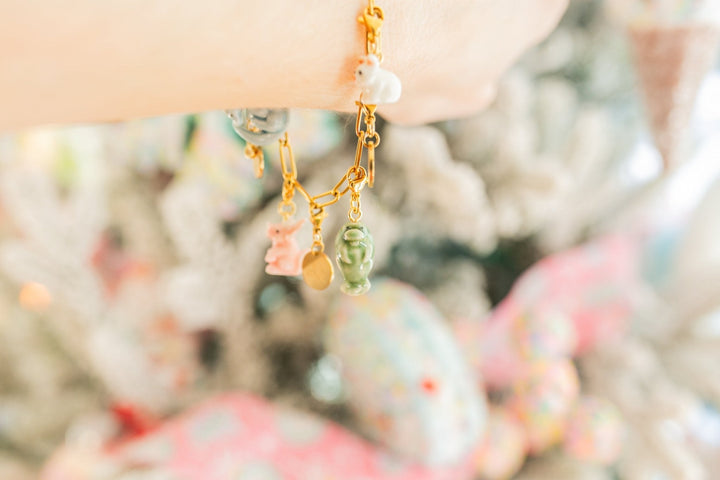 A close-up lifestyle shot of a delicate gold chain bracelet worn on a wrist, featuring pastel charms including a pink bunny, a green frog, and white pearls, set against a soft, blurred background.