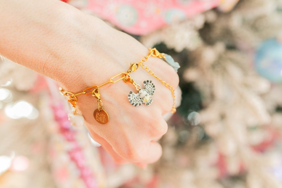 A close-up of a person's hand wearing a delicate gold charm bracelet with a heart-shaped charm and a silver-and-white patterned heart charm, set against a soft, blurred festive background.