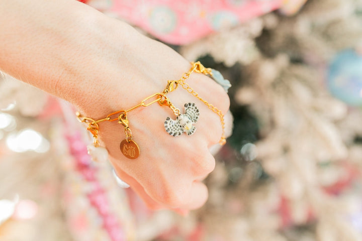A close-up of a person's hand wearing a delicate gold charm bracelet with a heart-shaped charm and a silver-and-white patterned heart charm, set against a soft, blurred festive background.