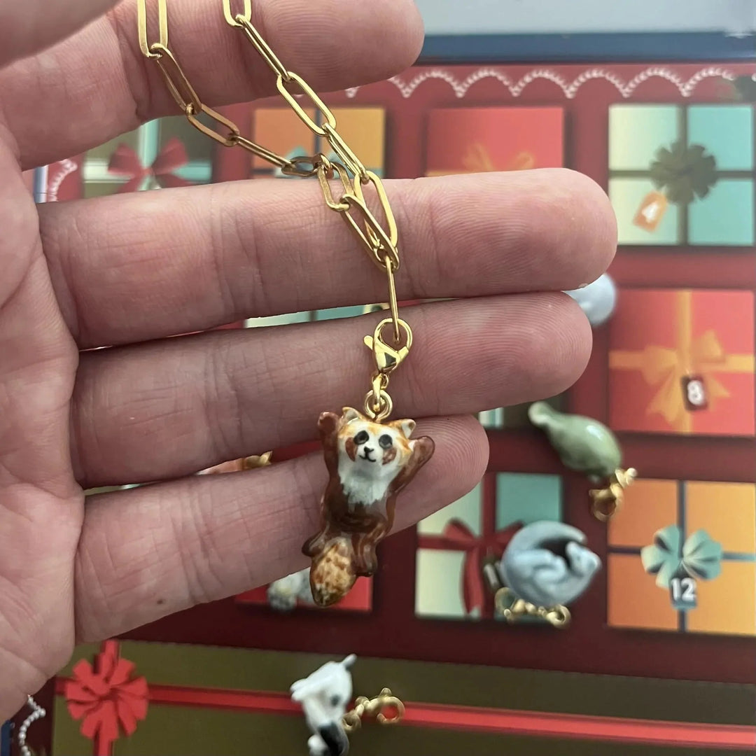 A hand holding a gold paperclip chain necklace with a small red panda pendant, with a Christmas advent calendar visible in the blurred background.