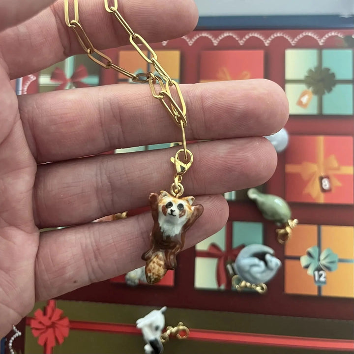 A hand holding a gold paperclip chain necklace with a small red panda pendant, with a Christmas advent calendar visible in the blurred background.