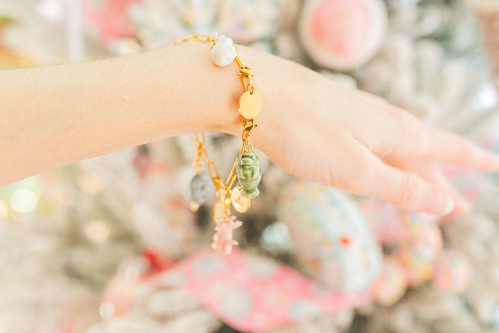A gold charm bracelet with pastel-colored beads and dangling charms worn on a person's wrist, set against a soft, blurred festive background.
