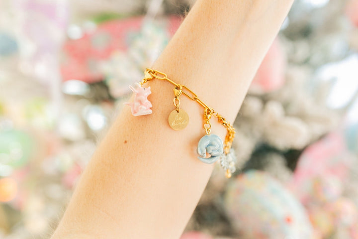 A gold charm bracelet worn on a person's wrist, featuring pastel pink, blue, and gold charms against a soft, colorful bokeh background.