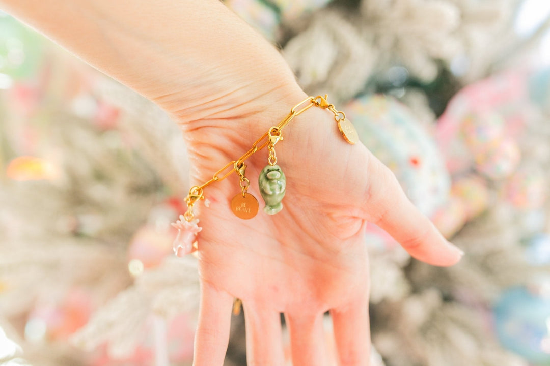 A gold charm bracelet with a green frog charm and small dangling charms, worn on a person's wrist against a soft, blurred festive background.