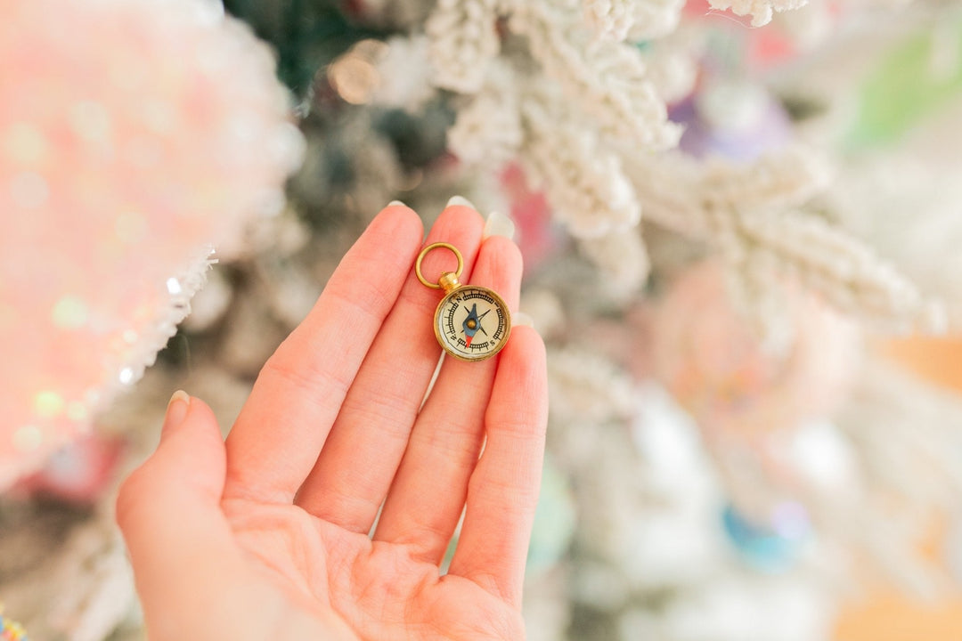 A hand holding a small gold compass pendant charm with a vintage-style illustration on a circular face, set against a soft, blurred festive background.