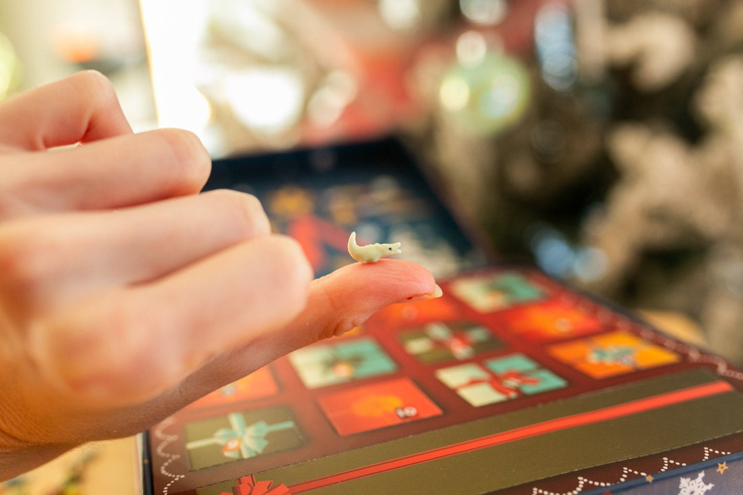 A close-up of a hand holding a tiny white mouse figurine over a colorful advent calendar box with festive holiday illustrations.