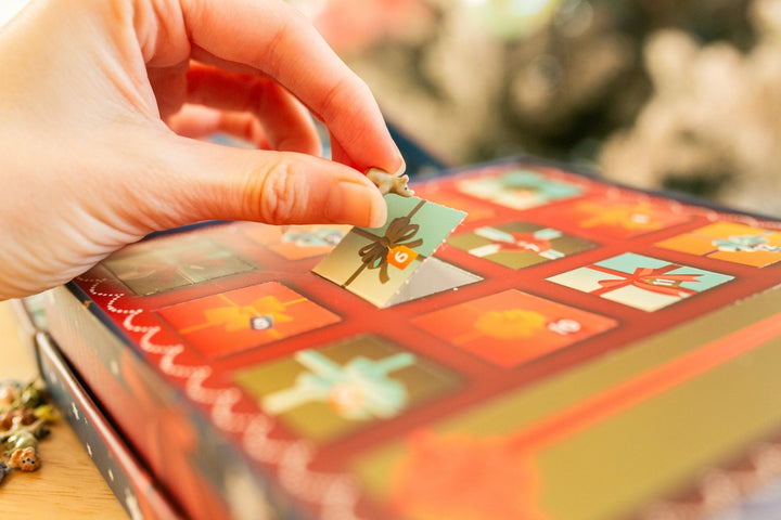 A close-up of a hand placing a small decorative tile onto a colorful holiday advent calendar box.