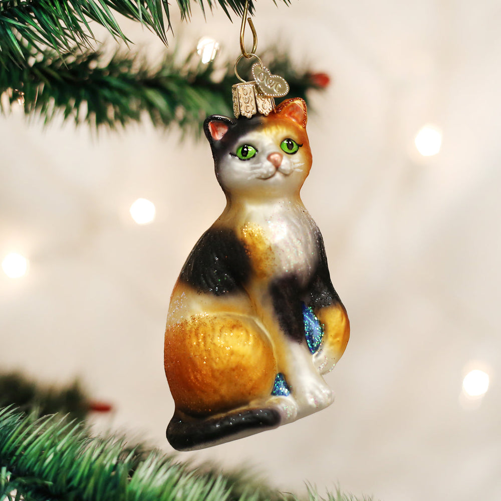 Calico cat ornament hanging on a Christmas tree with soft twinkle lights behind