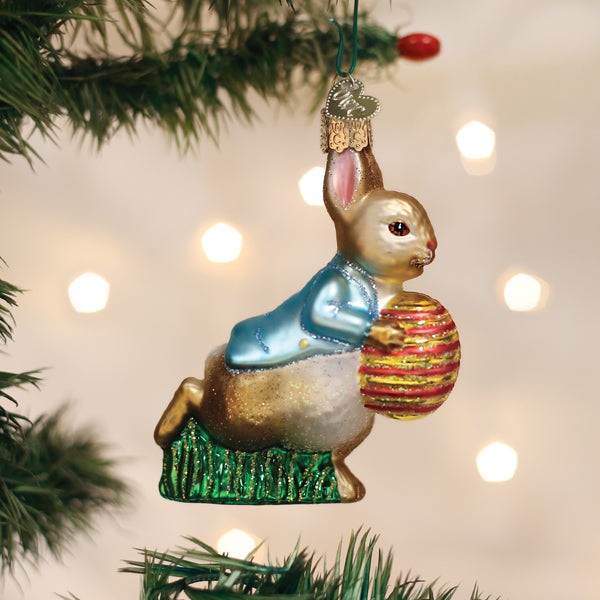 Peter Rabbit glass ornament in a blue jacket carrying a striped Easter egg, hanging on a Christmas tree