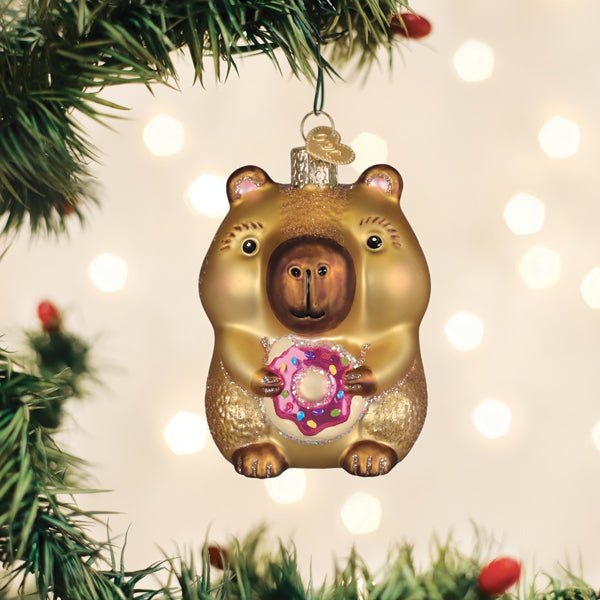 Capybara eating donut ornament hanging on a lit Christmas tree with warm bokeh lights