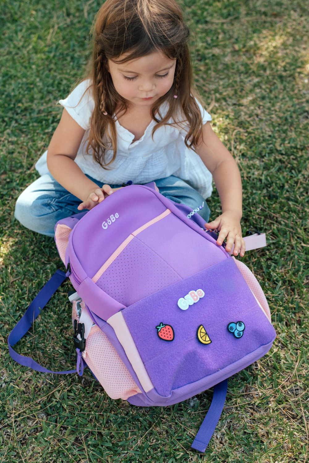 Child kneeling on grass beside a purple GoBe lunch bag clipped to a backpack