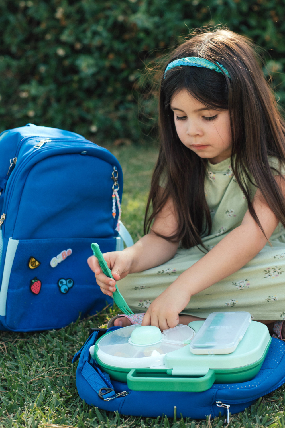 Girl on the grass unpacking an open lunchbox beside a blue backpack