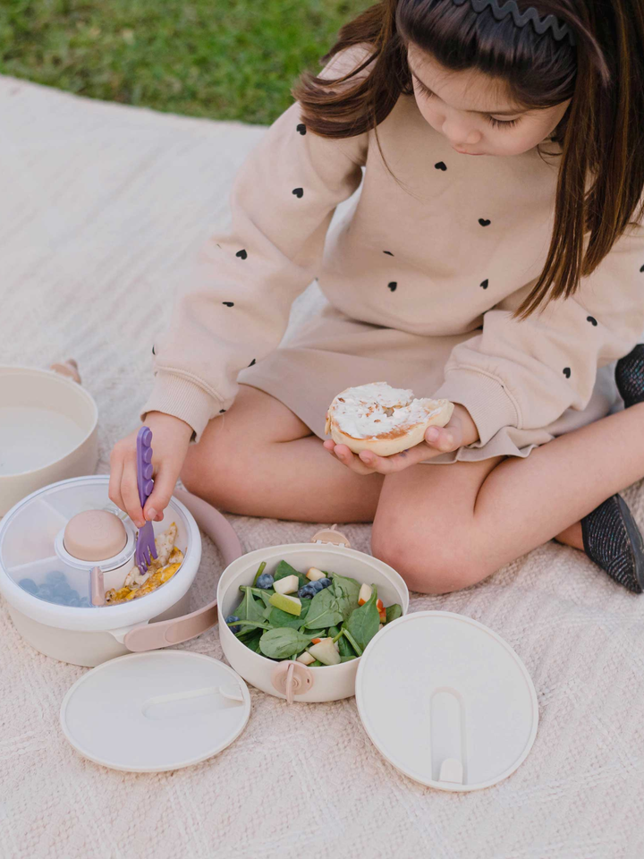 Girl eating a sandwich on a picnic blanket with the cream Lunch Stack open beside her