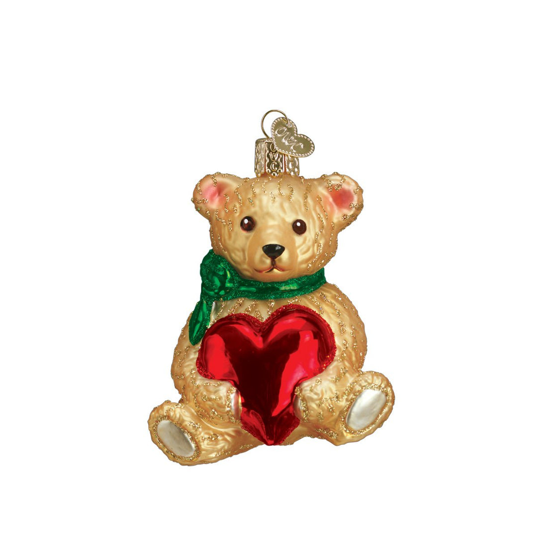 Seated honey teddy bear ornament in a green scarf hugging a shiny red heart, on white