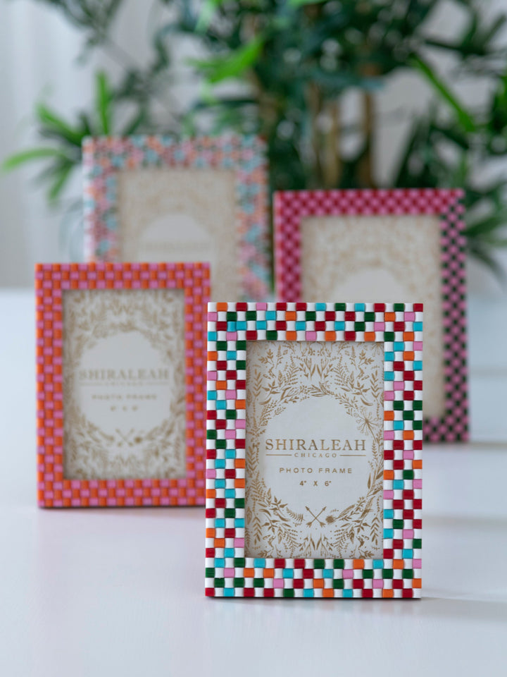 Four checkerboard picture frames grouped on a white shelf beside a green plant, in pink, red, and multicolor mosaic