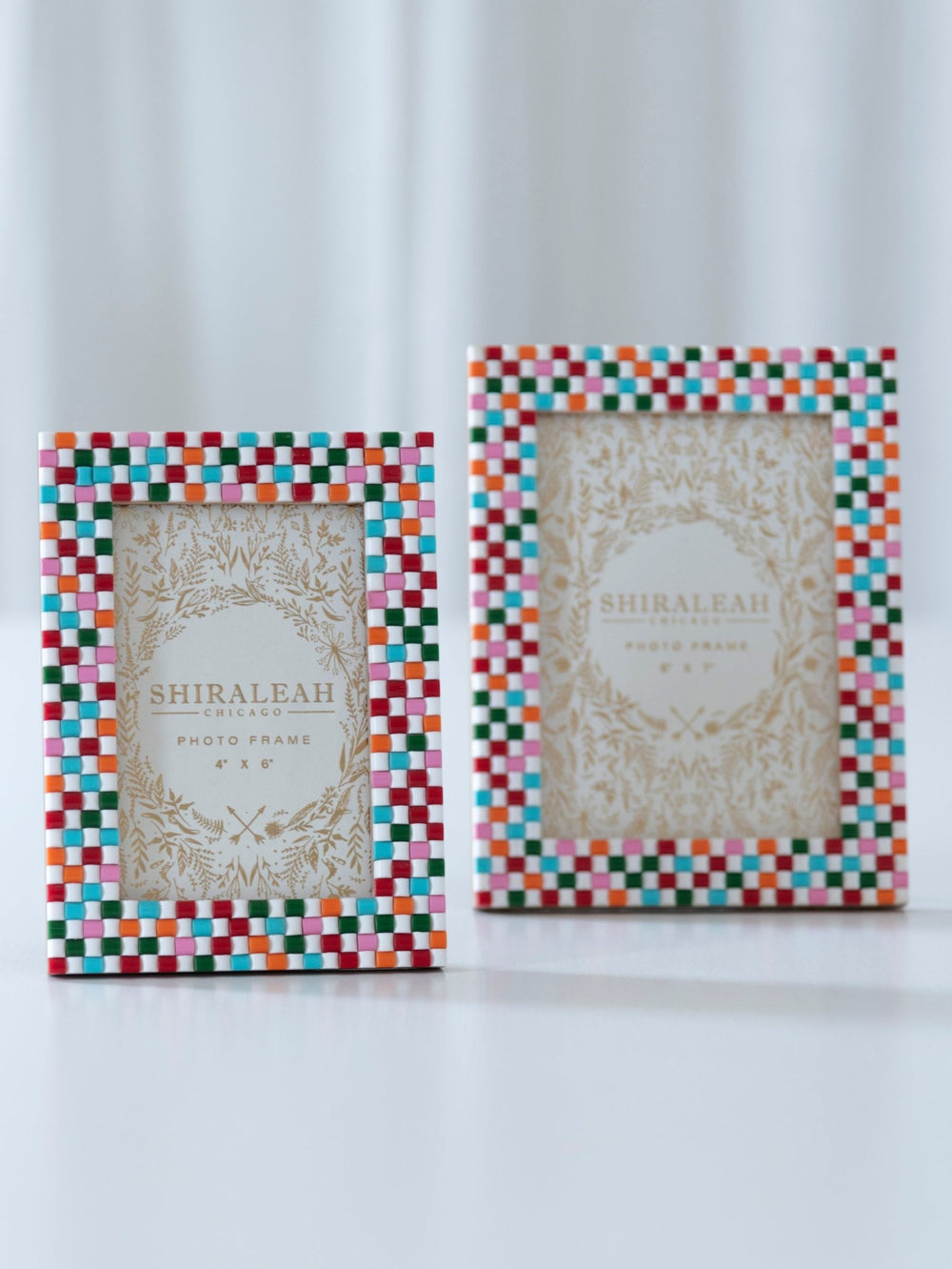 Two Shiraleah photo frames with colorful checkered borders, one small and one large, standing on a white surface against a soft white curtain background.