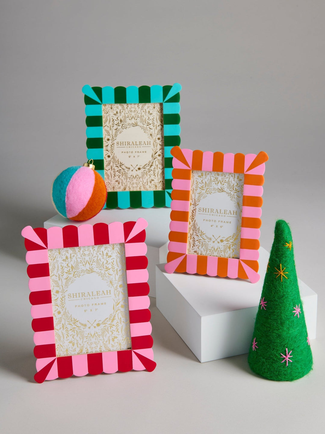Three decorative photo frames with striped felt edges in teal and green, pink and orange, and red and pink, displayed on white blocks with a small green felt tree and a striped ball prop against a light gray background.