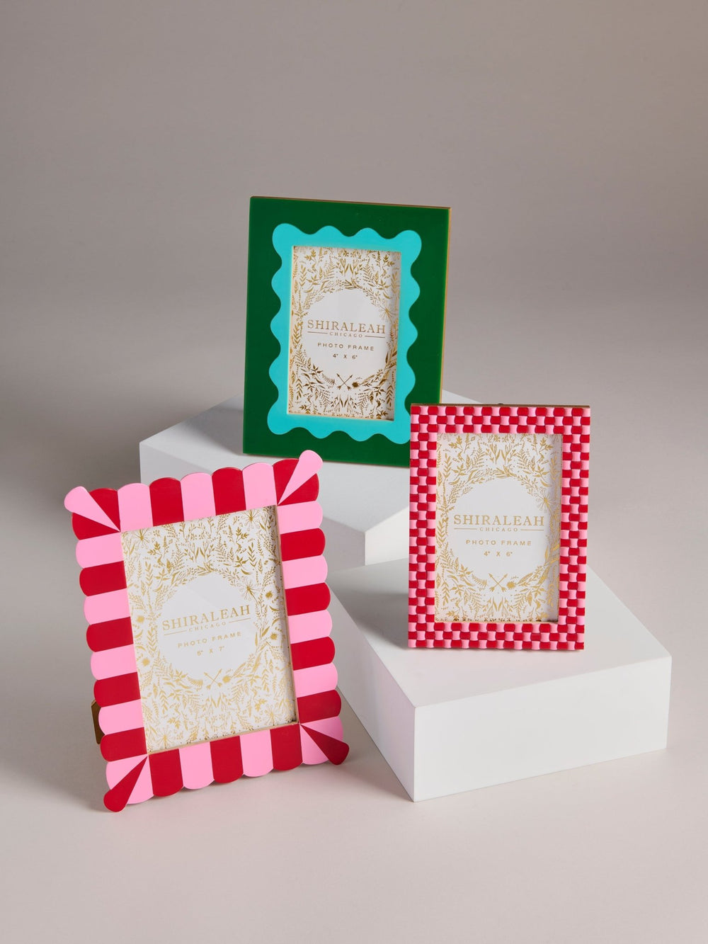 Three decorative picture frames displayed on white pedestals against a neutral studio background. One frame has a green border with a turquoise inner border, one has a pink and red striped border, and one has a red and white checkered border.