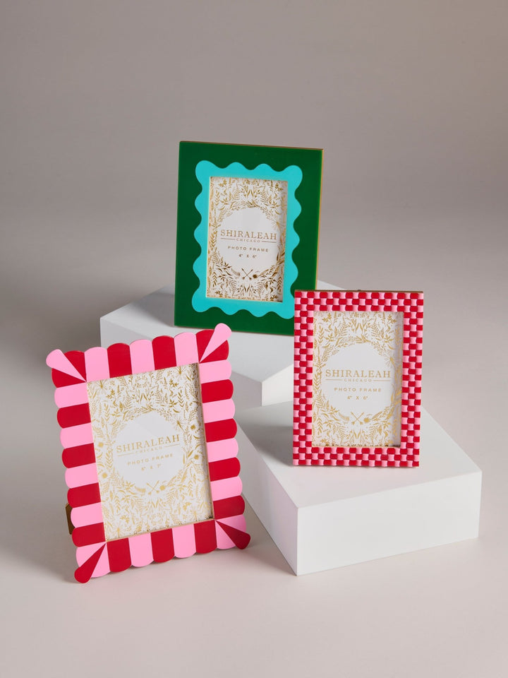 Three decorative picture frames displayed on white pedestals against a neutral studio background. One frame has a green border with a turquoise inner border, one has a pink and red striped border, and one has a red and white checkered border.