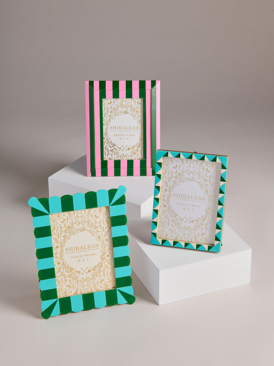 Three decorative picture frames displayed on white pedestals against a neutral studio background. The frames feature colorful striped and geometric borders in green, turquoise, pink, and cream with ornate gold detailing in the center.
