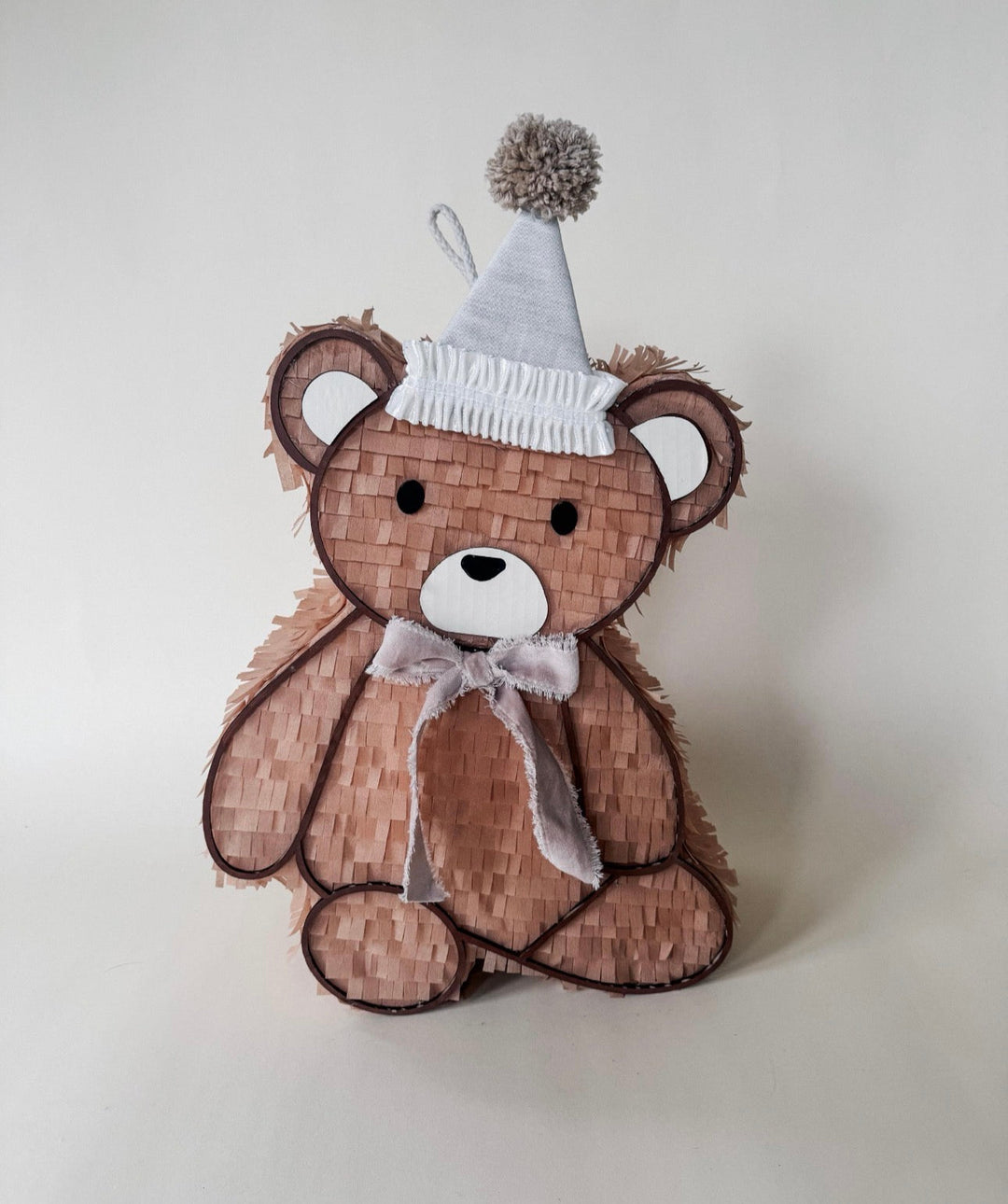 Light brown teddy bear piñata with a white fringed party hat, gray pom-pom, and tied ribbon bow on a plain backdrop
