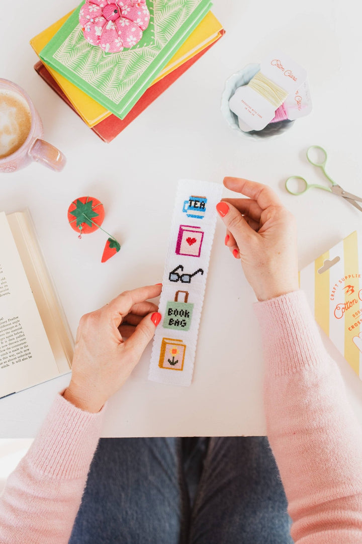 Bookish cross stitch bookmark draped over an open book with craft supplies