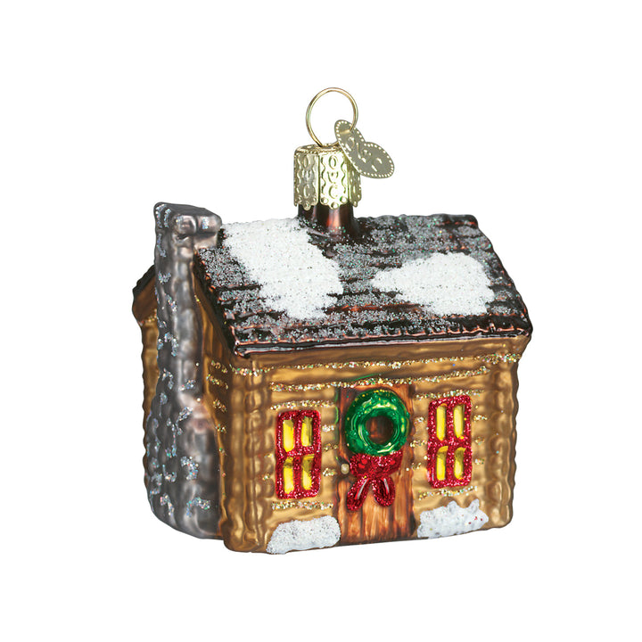 Brown log cabin glass ornament with snowy roof, glowing windows, and wreath on red door, on white