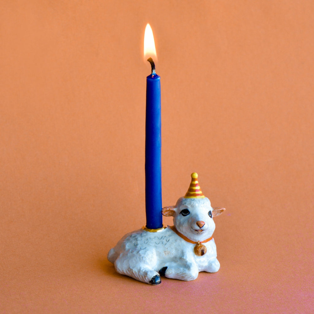 Year of the Goat porcelain lamb candle holder in a party hat holding a lit blue taper candle for a birthday cake