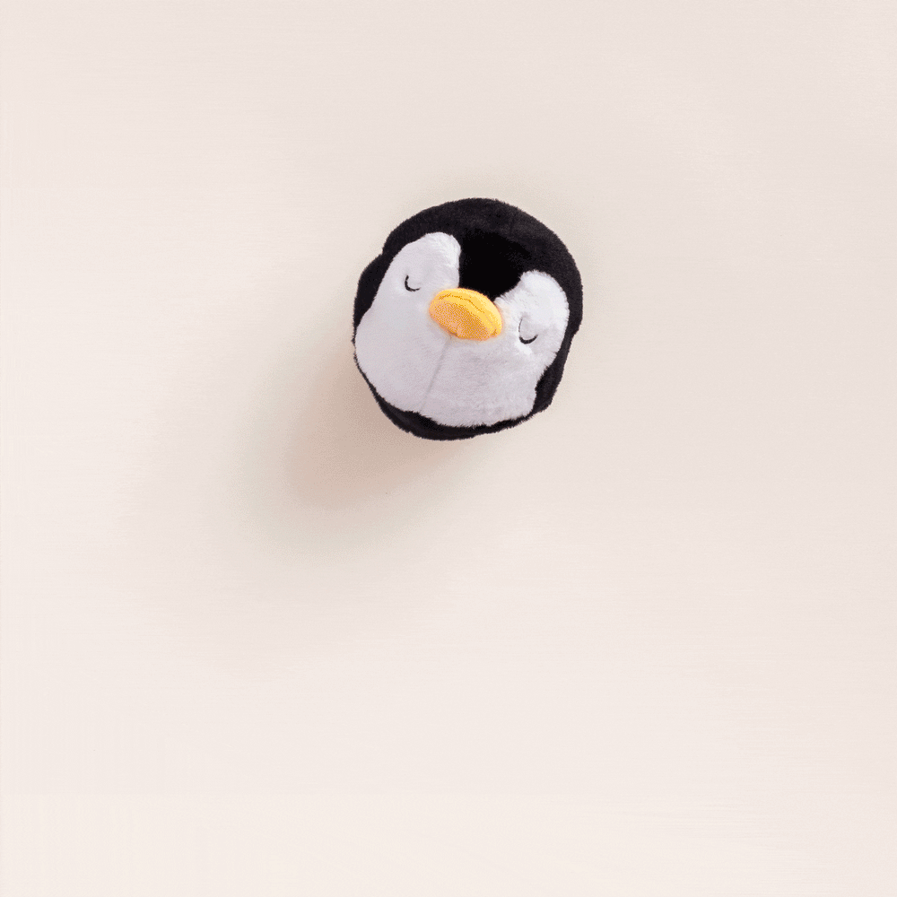 Penguin plush dog toy seen from above on a pale pink background.