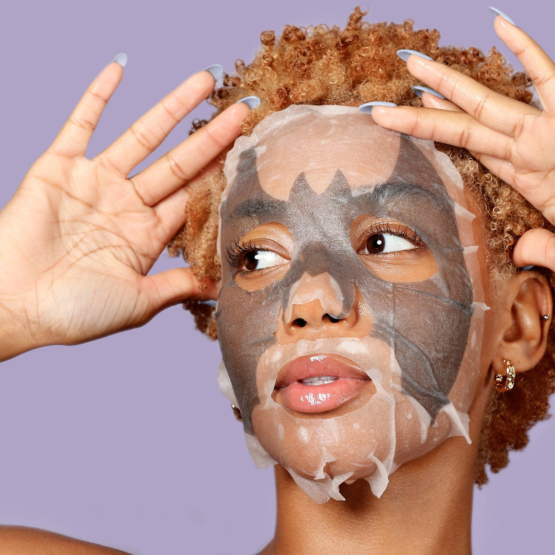 Model in the bat design charcoal sheet mask glancing sideways with hands raised, playful Halloween party pampering