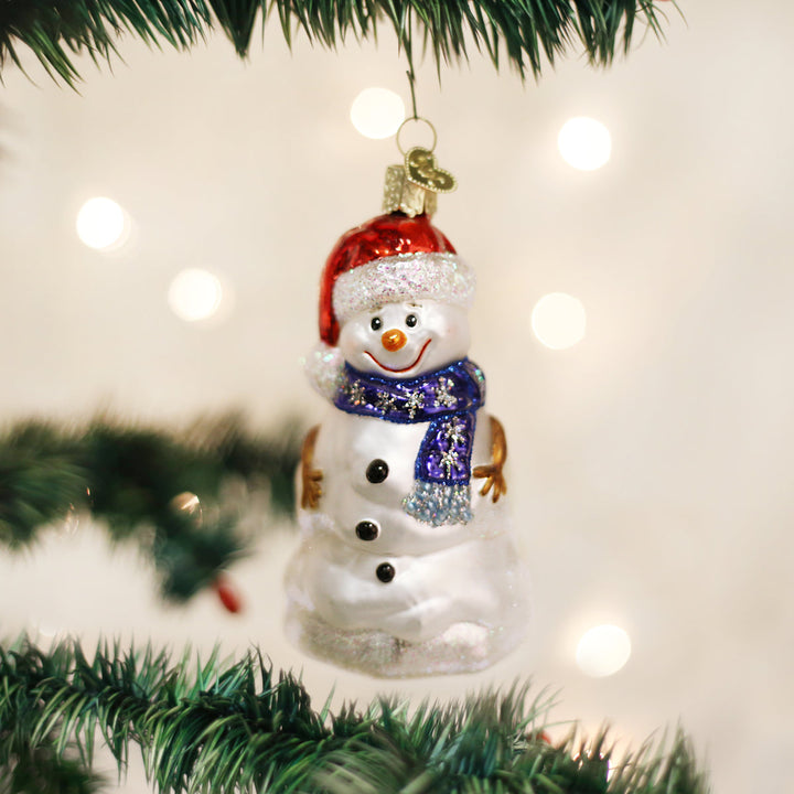 Happy snowman glass ornament with twig arms hanging on a lit Christmas tree