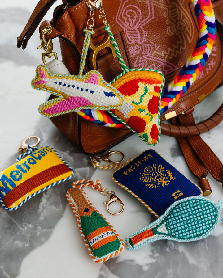 Needlepoint bag charms including an airplane, pizza slice, subway card, champagne, and passport clipped to a leather bag
