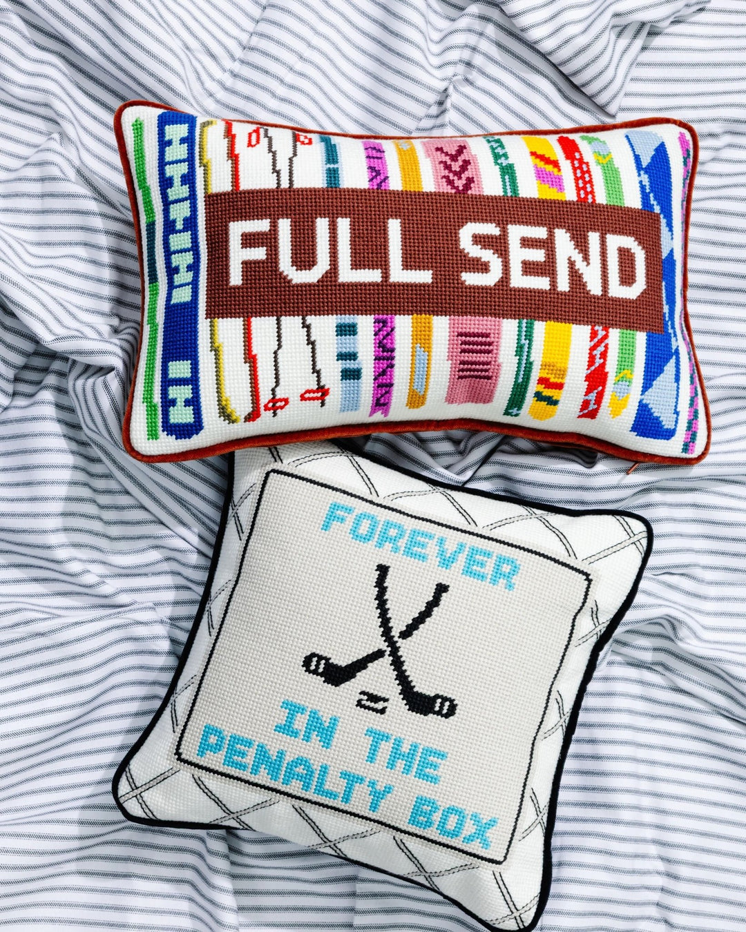 Full Send ski needlepoint pillow and a Forever in the Penalty Box hockey needlepoint pillow styled on striped bedding