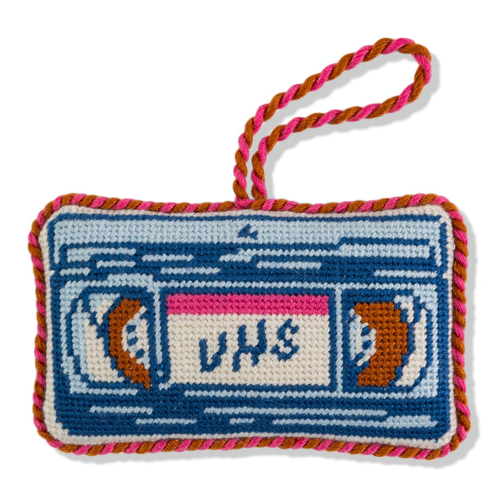VHS tape needlepoint ornament, front view of a blue cassette with white label and red and white cord