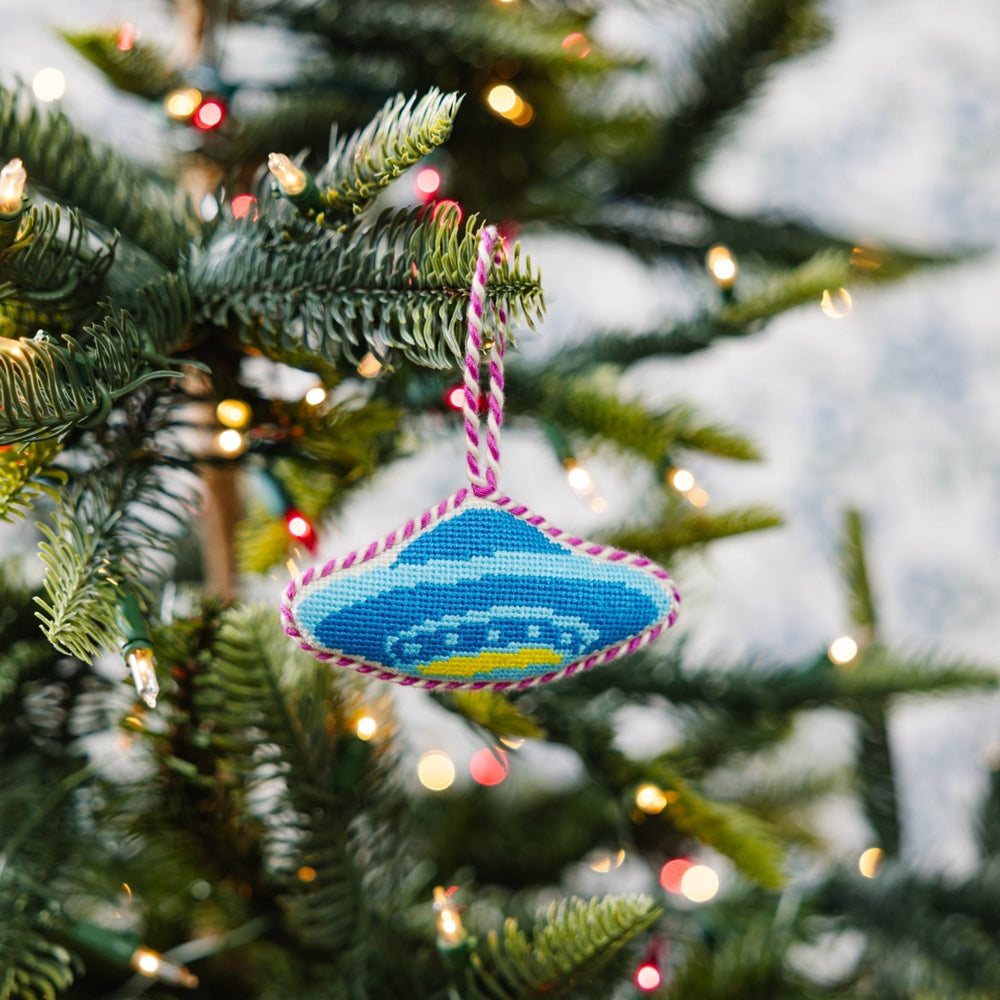 UFO needlepoint ornament hanging on a Christmas tree branch with soft string lights behind