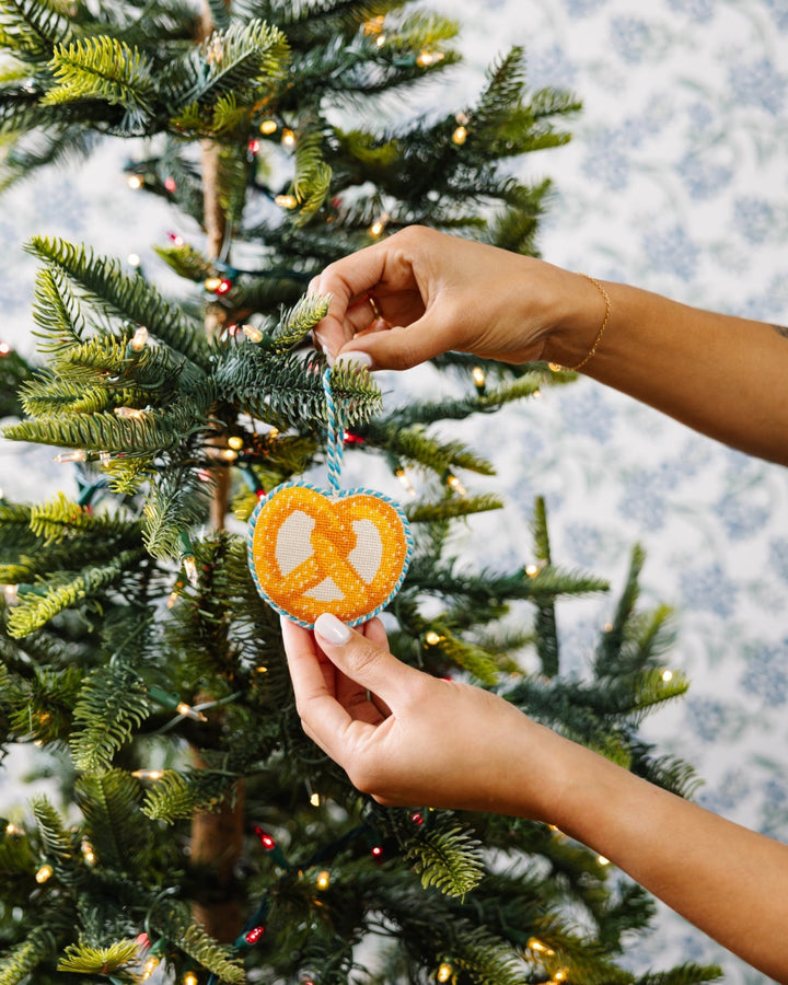 Two hands hanging the pretzel needlepoint ornament on a tree against blue toile wallpaper
