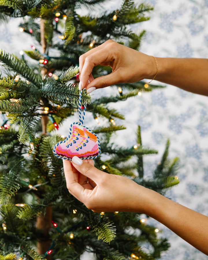 Two hands placing the pink snow boots needlepoint ornament on a Christmas tree