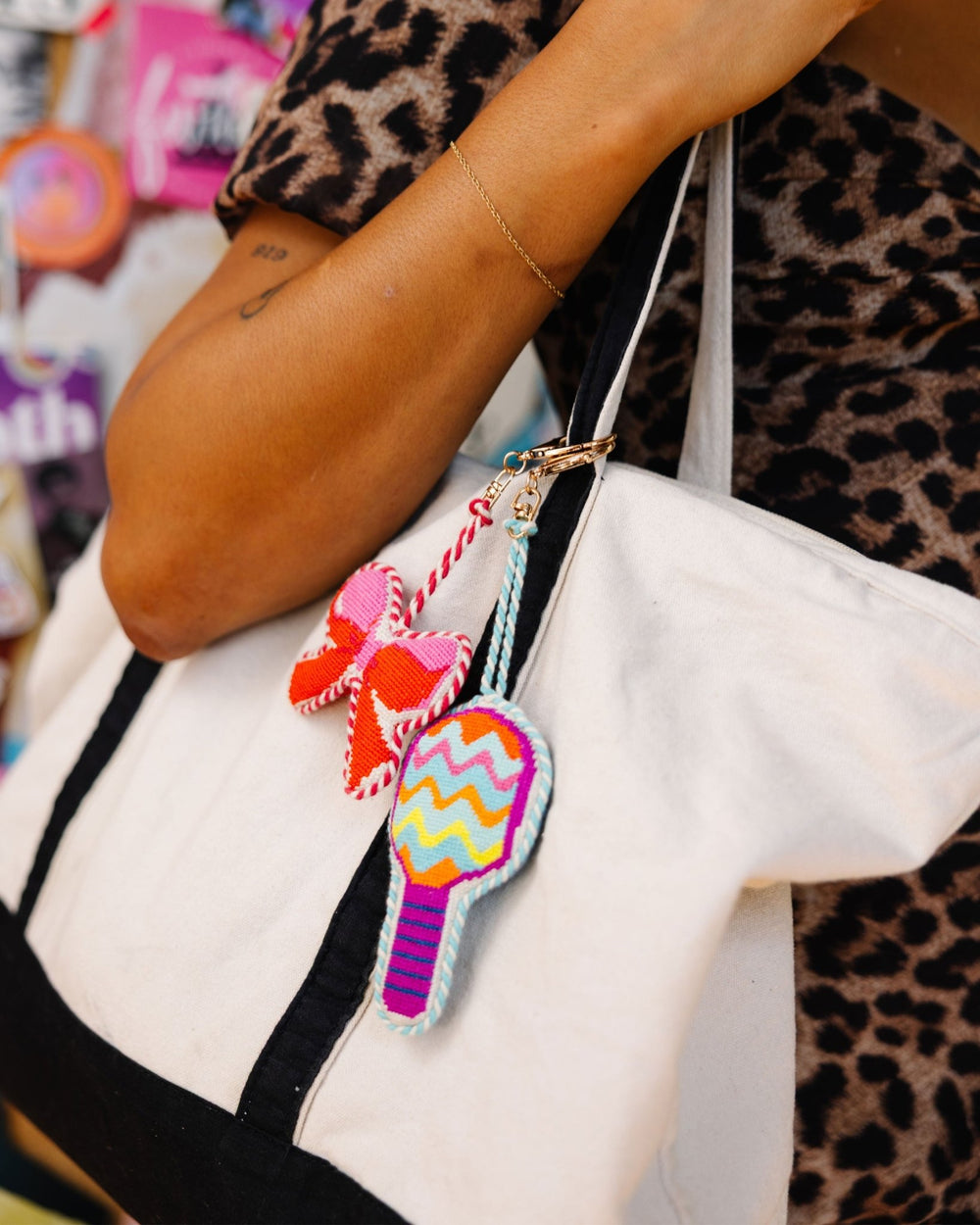 Canvas tote bag styled with a pink bow needlepoint charm and a colorful chevron bag charm held by a person in leopard print
