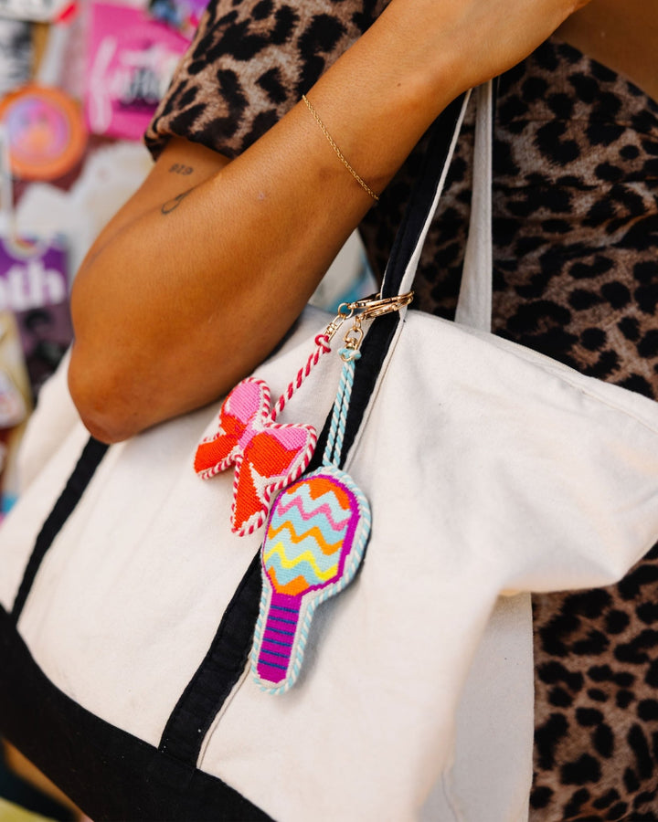 Canvas tote bag styled with a pink bow needlepoint charm and a colorful chevron bag charm held by a person in leopard print