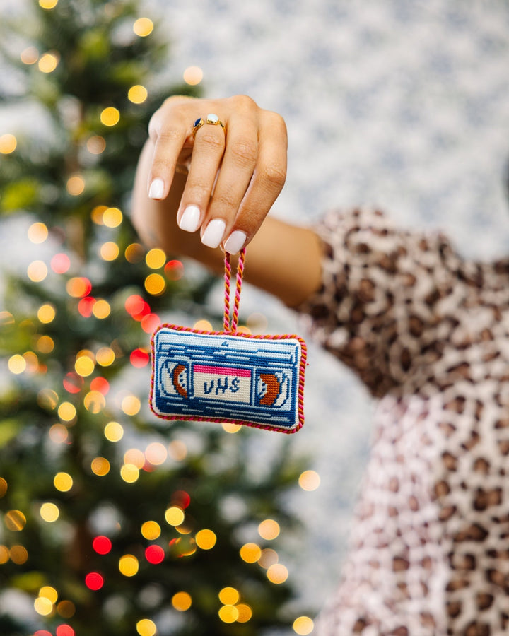 Hand in a leopard print sleeve holding the VHS needlepoint ornament in front of a lit tree