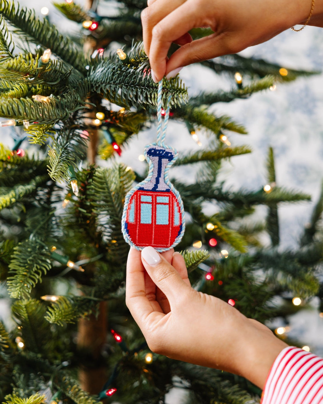 Two hands hanging the ski lift gondola needlepoint ornament on a Christmas tree