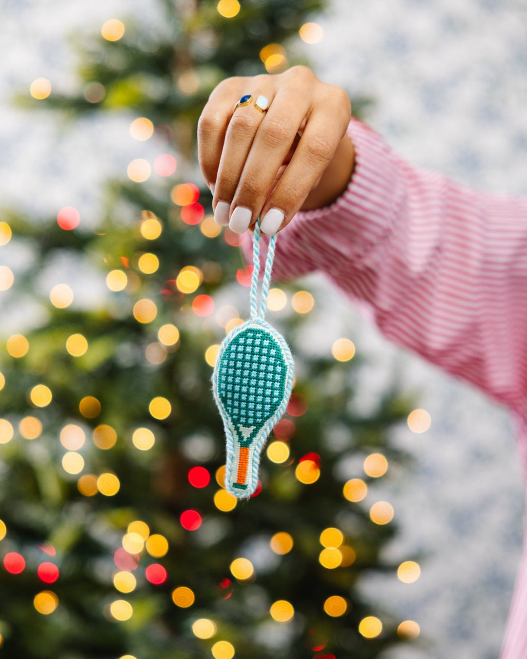 Hand in a pink striped sleeve holding the tennis racket ornament by its loop near a lit tree