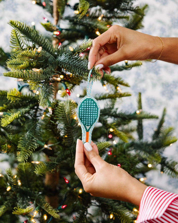 Two hands hanging the tennis racket needlepoint ornament on a tree against blue toile wallpaper
