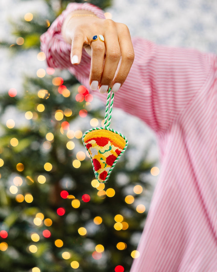 Hand in a pink striped sleeve holding the pizza ornament by its loop in front of a lit tree