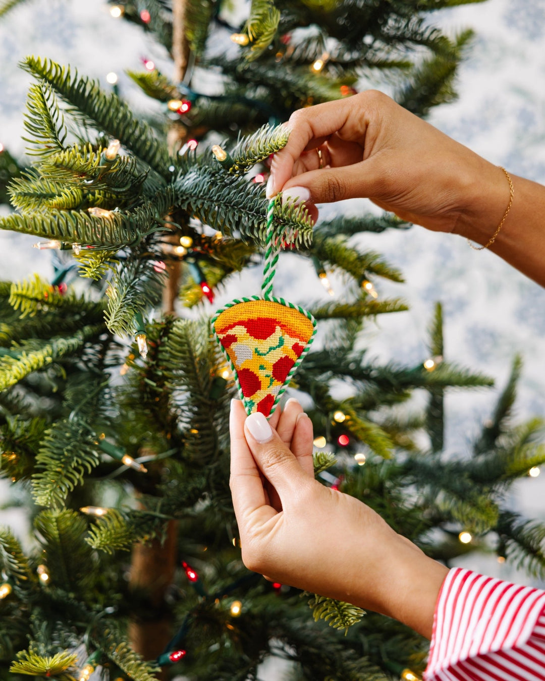 Two hands hanging the pizza slice needlepoint ornament on a Christmas tree