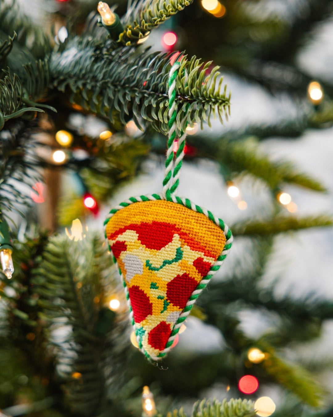 Pizza ornament hanging from a green Christmas tree branch with soft twinkle lights behind it