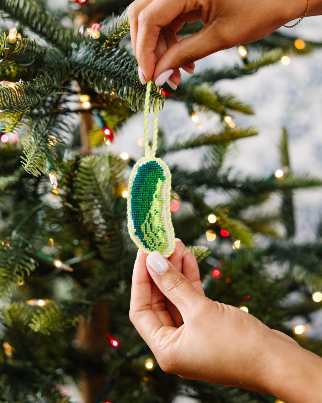 Two hands hanging the green pickle ornament on a tree in front of blue toile wallpaper