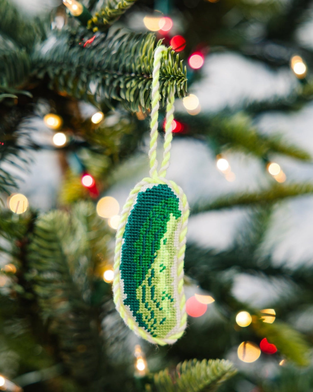 Pickle needlepoint ornament hanging from a Christmas tree branch with string lights behind
